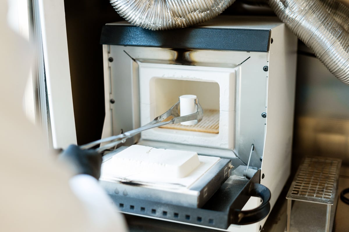 A technician using tongs to place a cylindrical test sample into a small high-temperature laboratory furnace.