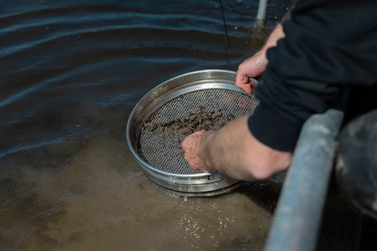 Hands passing material through a test sieve during sample preparation and sizing work.