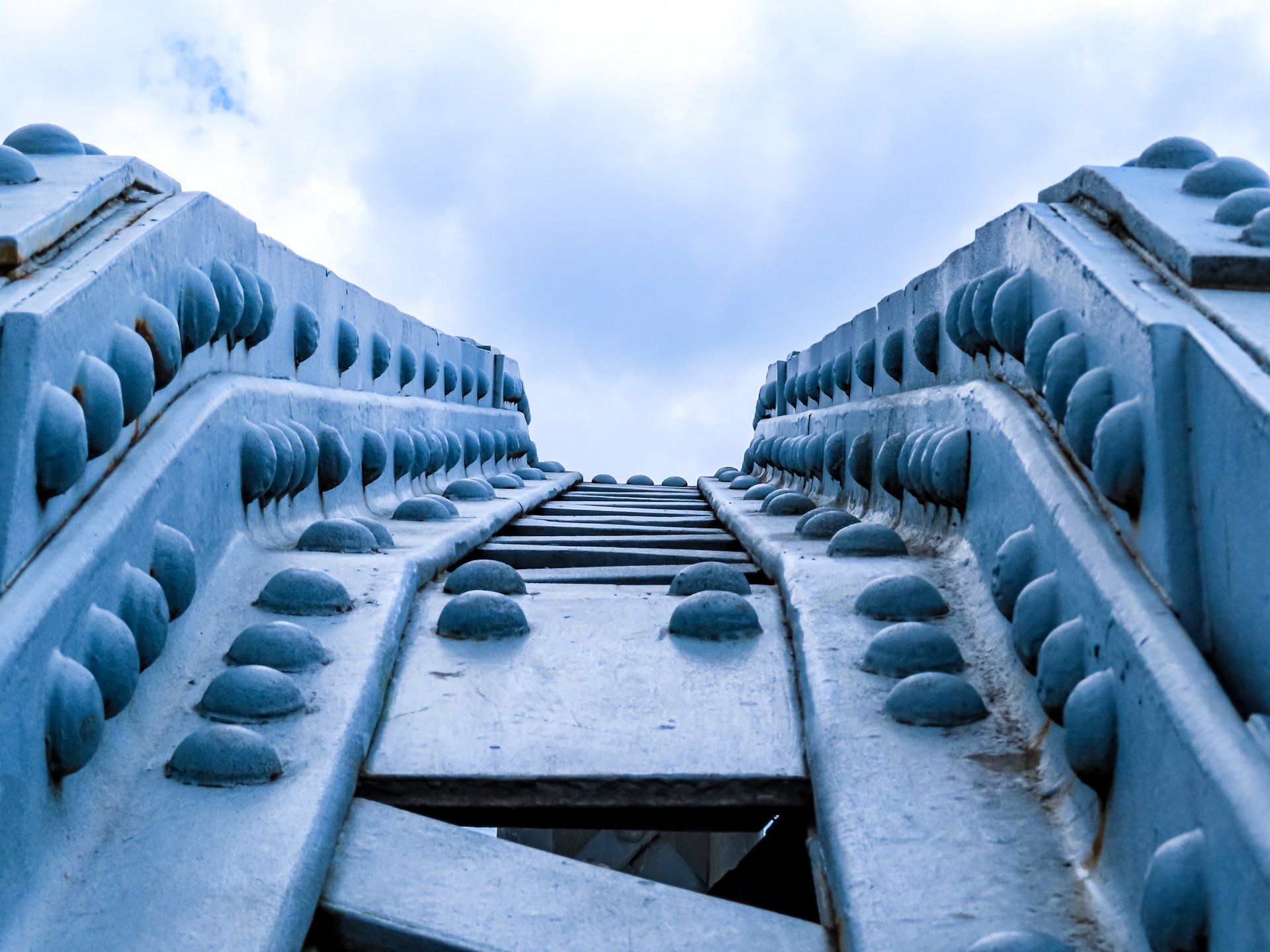 Low-angle view of a steel bridge framework with riveted connections against a cloudy sky