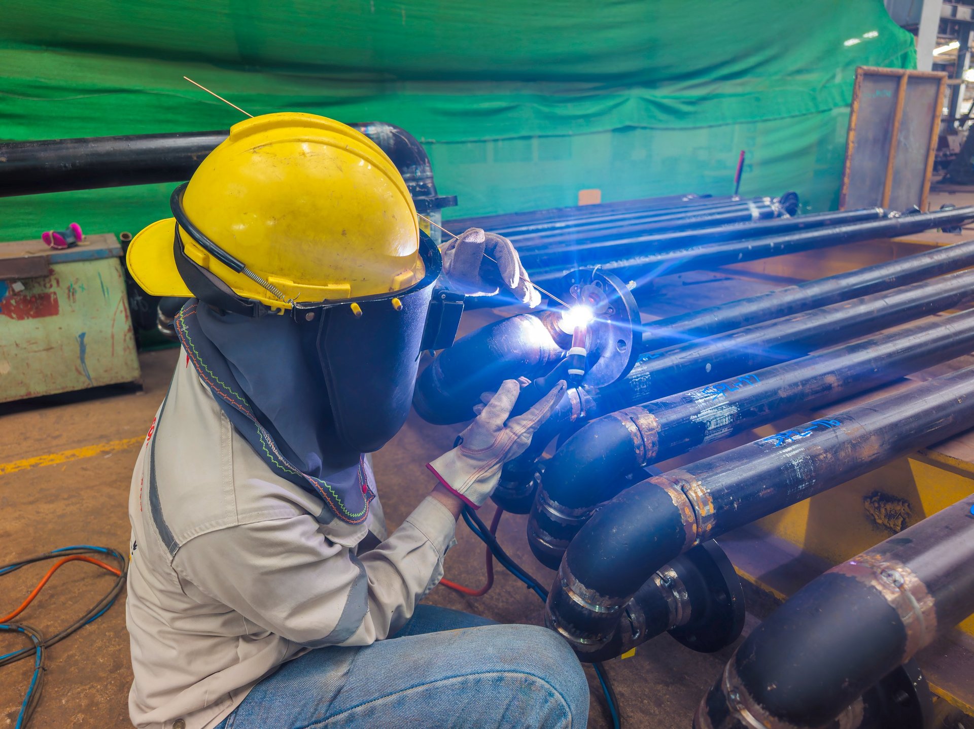 Welder in a safety helmet using a torch to weld a steel pipe flange inside an industrial pipe workshop, bright arc light