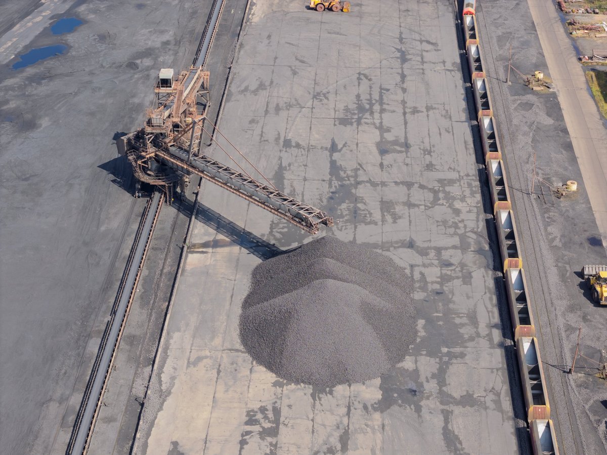Bulk material being stacked into a conical stockpile at a shipping yard with rail wagons alongside.