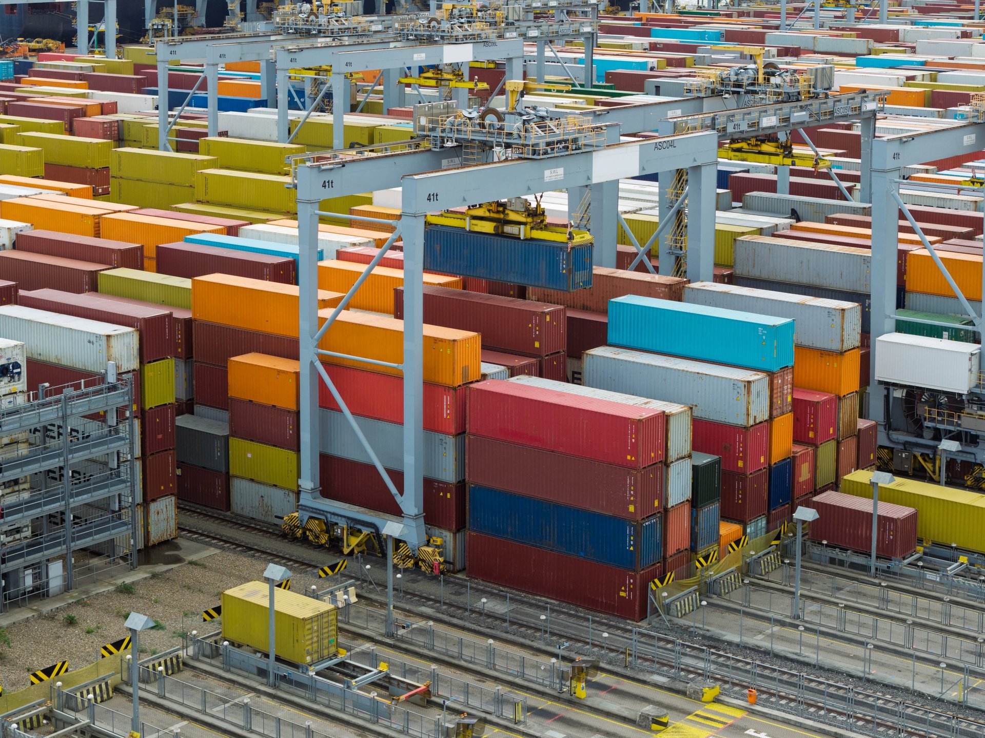 Gantry crane lifting a shipping container above stacked cargo at a port terminal